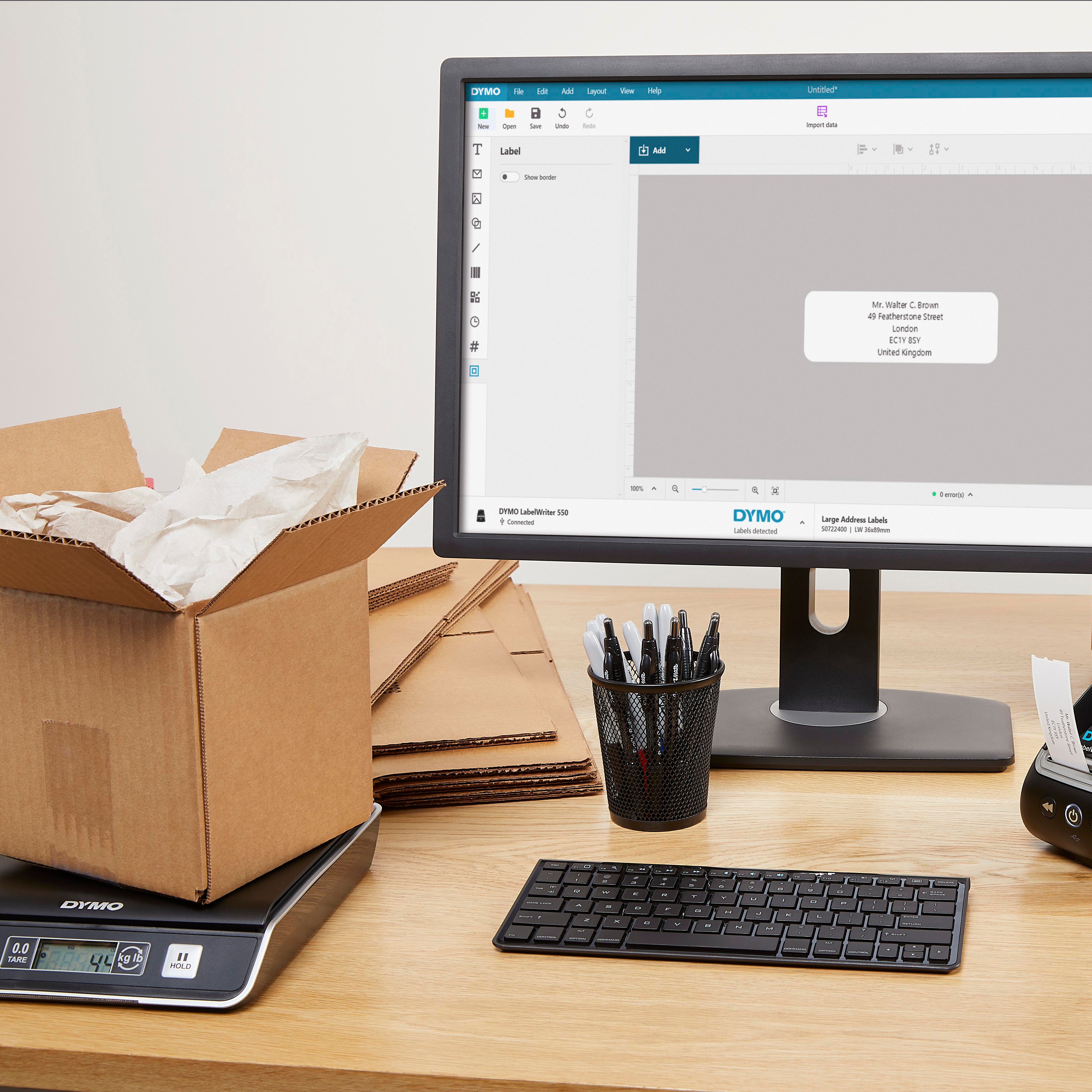 Packing workstation with DYMO postal scale, shipping box and computer screen showing address label design software for parcel processing