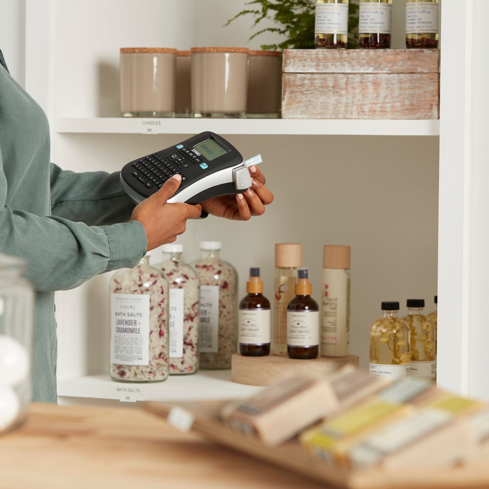 Person using a DYMO desktop label maker to organise and label candles, bath salts and beauty products on retail display shelves