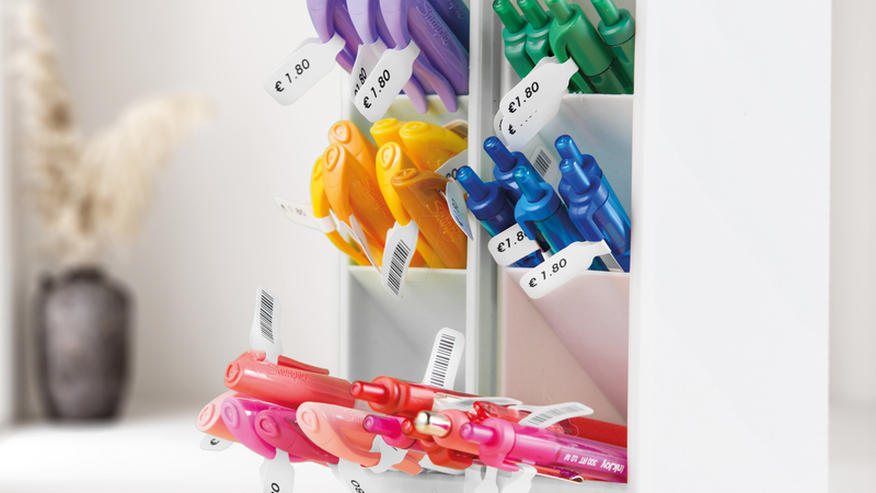 Colourful pens labeled with DYMO price tags displaying price in a retail pen holder. The small DYMO labels include both price and barcode, highlighting organised and professional labelling for office supplies or retail products.