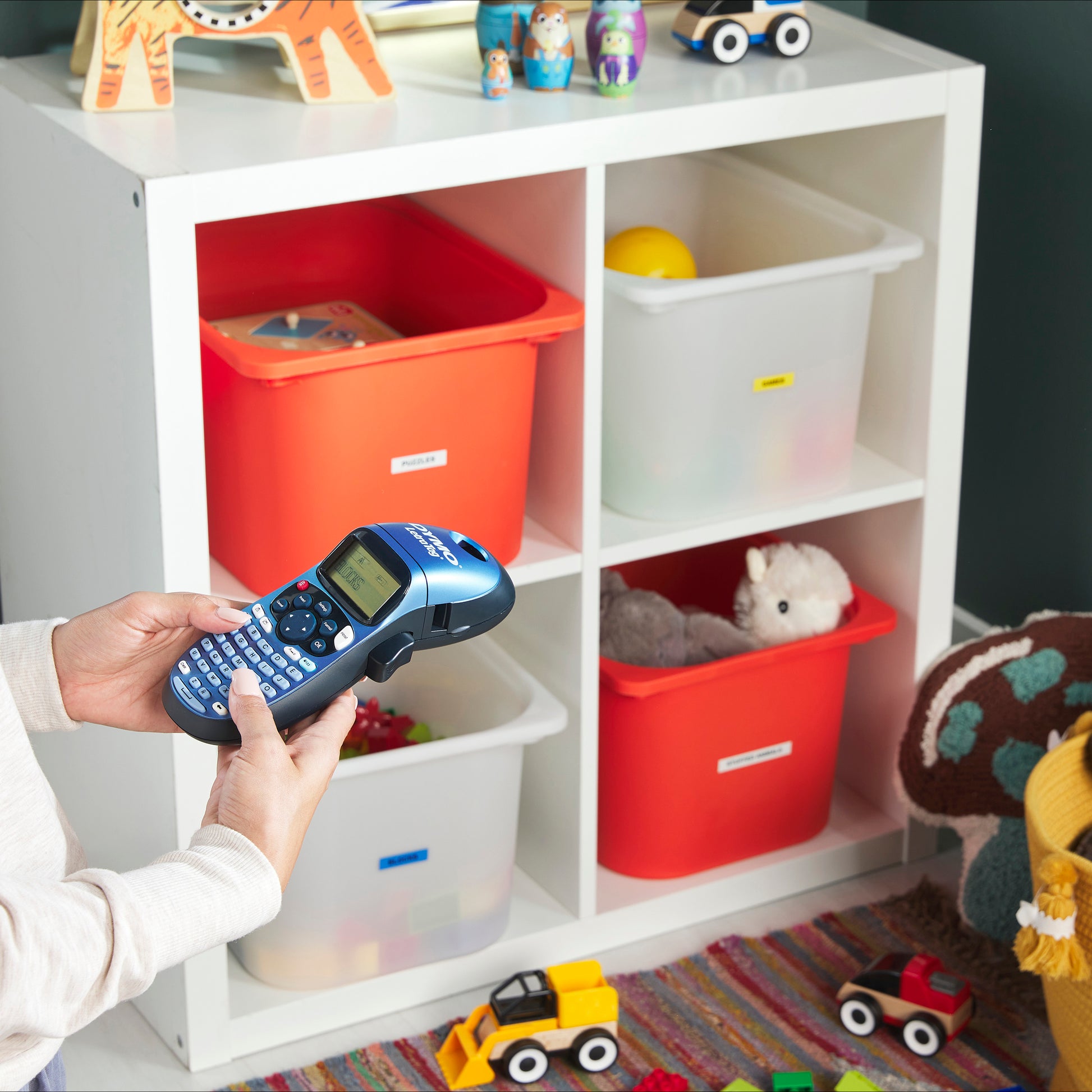 Person using a DYMO LetraTag label maker to organise labelled toy storage tubs in a playroom or classroom