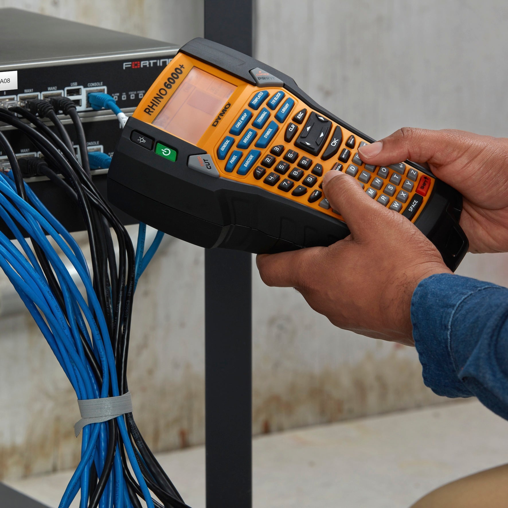 Technician using a DYMO Rhino 6000 Plus label printer to identify network cables beside rack mounted IT equipment
