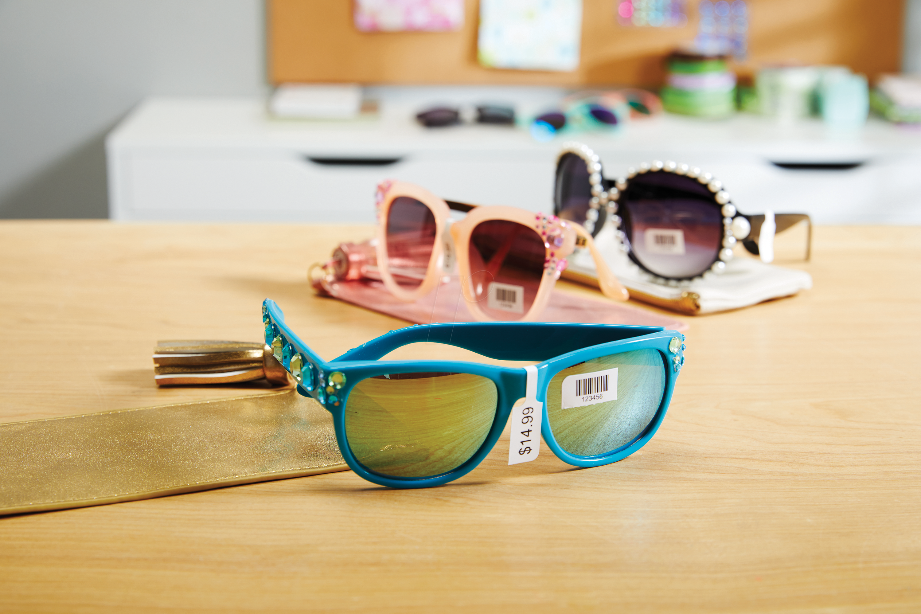Three pairs of colourful sunglasses displayed on a wooden surface, each labeled with a DYMO price tag, including a $14.99 tag on the front pair. The labels also include barcodes, showcasing the use of DYMO labels for clear and professional pricing in retail environments.