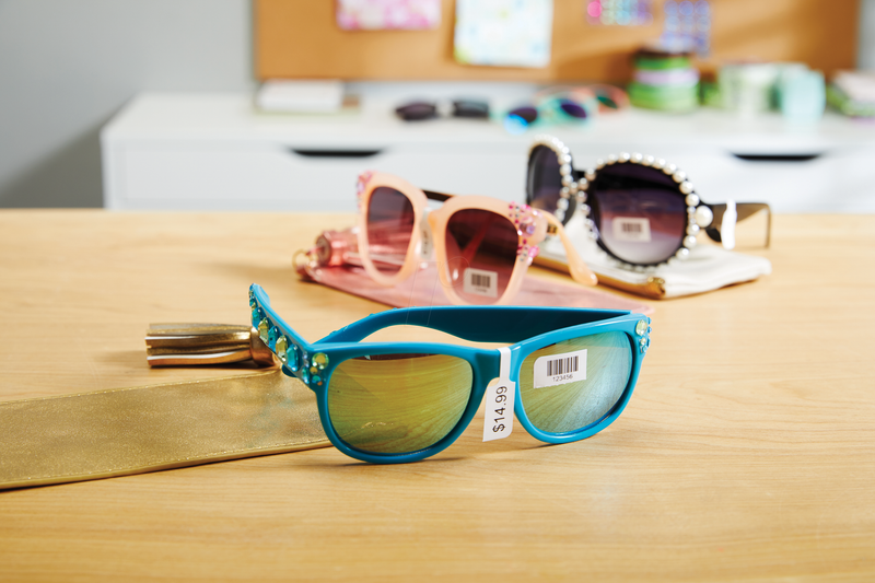 Three pairs of colourful sunglasses displayed on a wooden surface, each labeled with a DYMO price tag, including a $14.99 tag on the front pair. The labels also include barcodes, showcasing the use of DYMO labels for clear and professional pricing in retail environments.